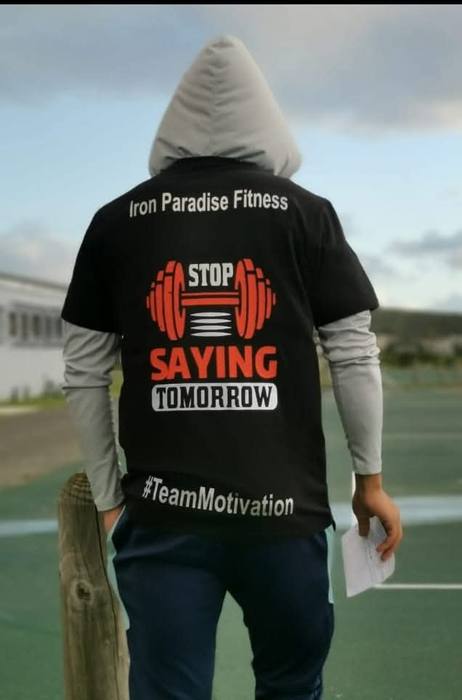 Back of a black gym t-shirt printed for Iron Paradise Fitness, reading Stop Saying Tomorrow above a red dumbbell